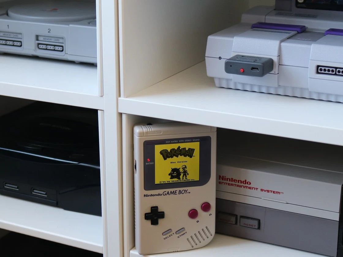A Game Boy beside the NES, with more consoles on our shelves