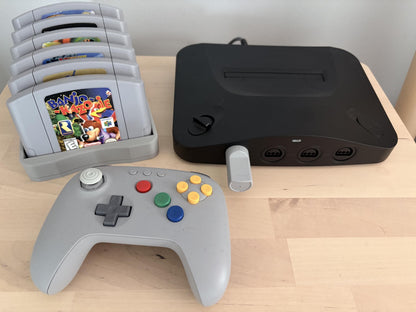 Gray six-game cartridge holder beside a console and controller on a wooden shelf; electronics and games are photo props.