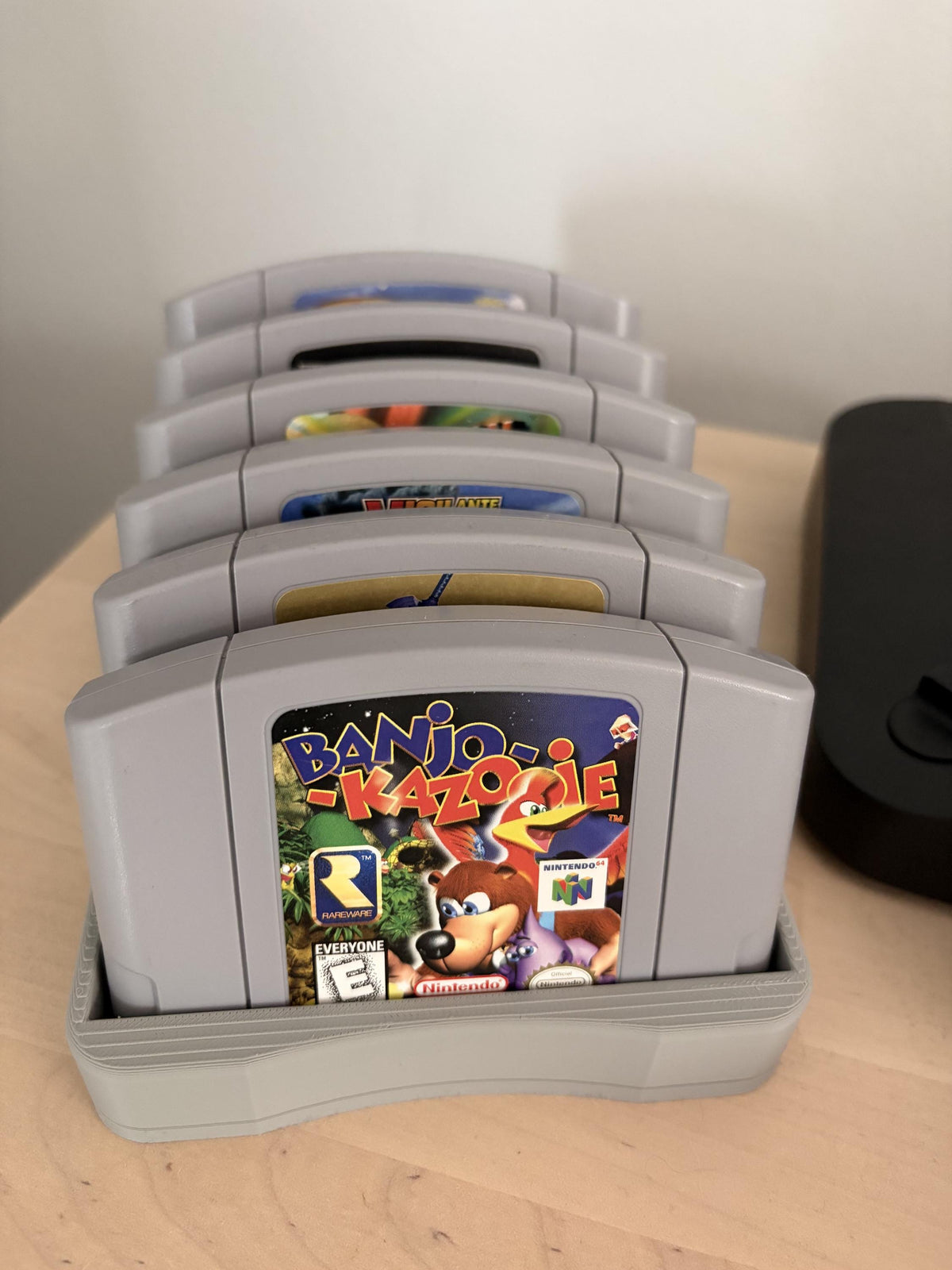 Gray holder with six Nintendo 64 cartridges displayed on a wooden shelf, viewed from the front.